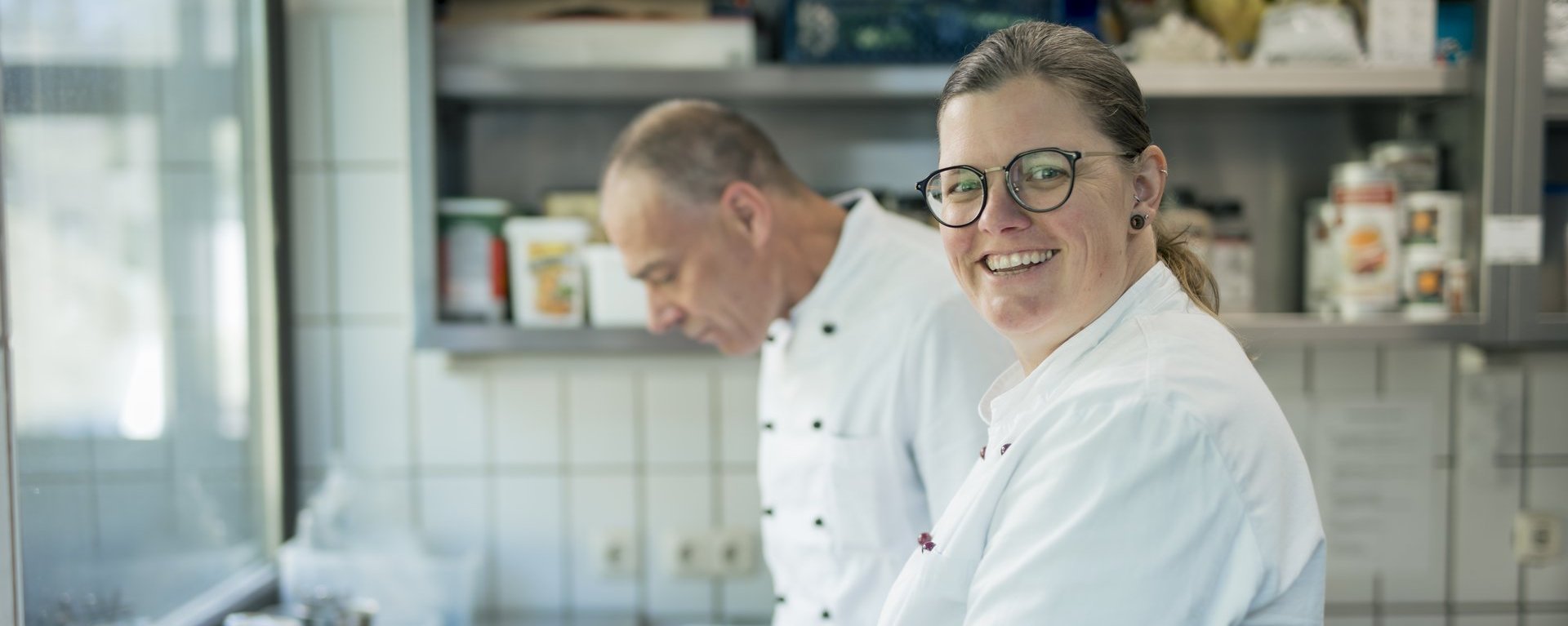 Eine Frau lächelt in die Kamera, während sie mit einem Mann in der Küche arbeitet.